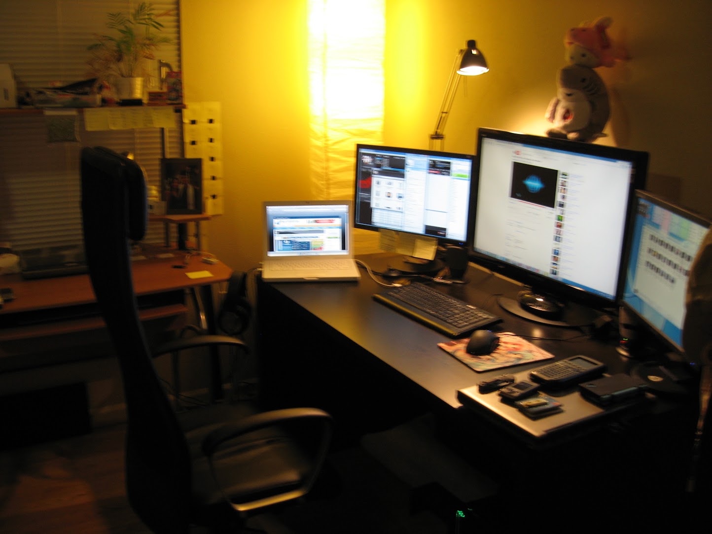 A dim home office around 2010: a black desk with a white MacBook and three external monitors showing poker tables and forums, a trackball mouse, and a row of retired cell phones.