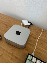 Apple Mac mini sitting on a wooden desk with a power cable plugged in. Compact, silver, about the size of a paperback.